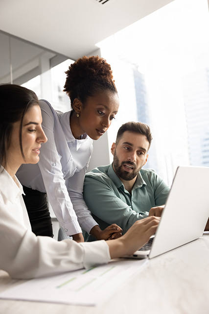 Multiethnic team of focused young managers working on Internet project together, using laptop at office table, discussing online sales strategy, pointing at screen, talking. Vertical shot